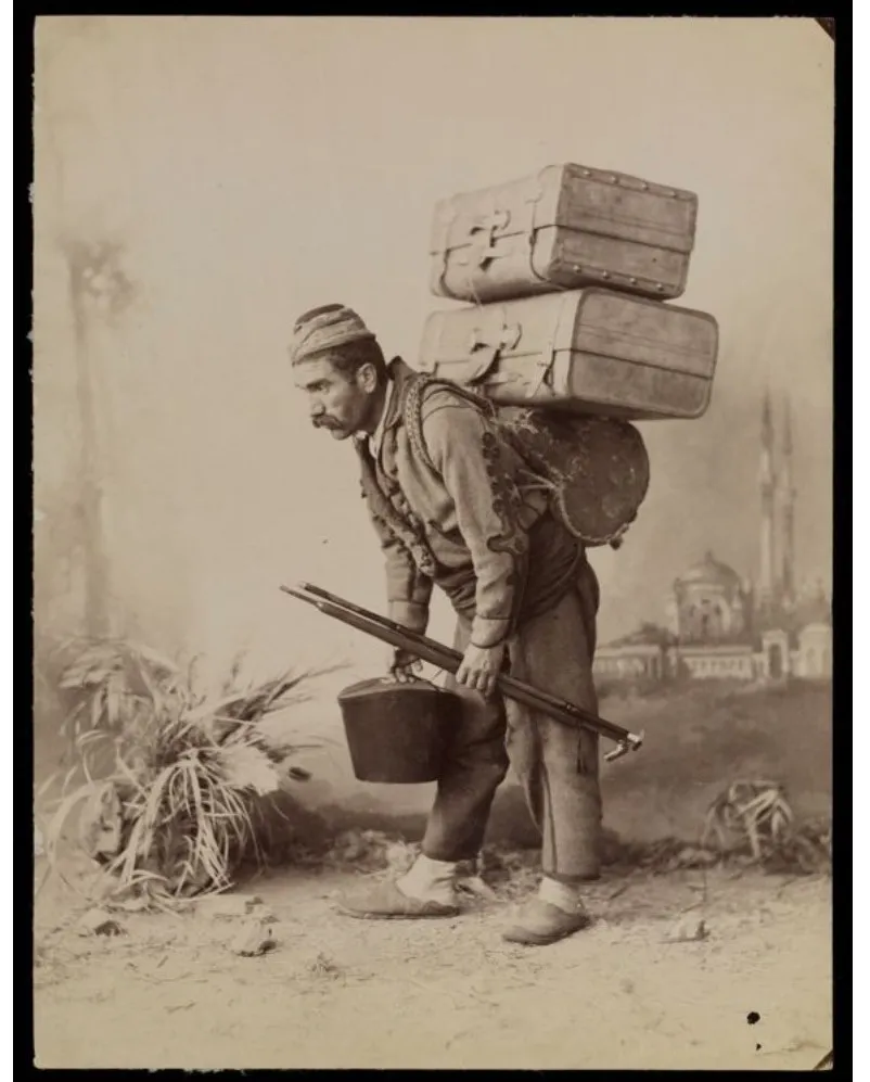Sepia studio photograph of an Ottoman-era porter bent under two strapped trunks, a kettle in one hand and a staff in the other, a painted mosque behind him