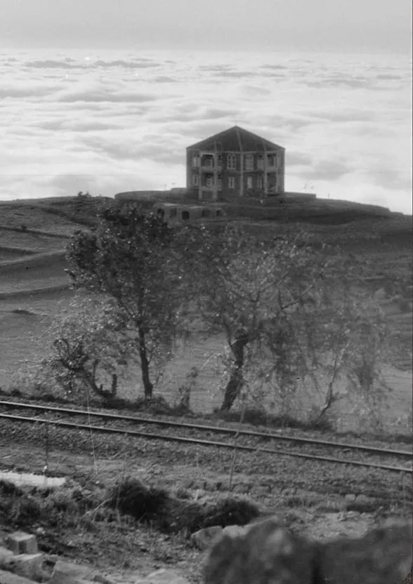Black-and-white photograph of a stone house on a hill above a railway line, cloud filling the valley behind.