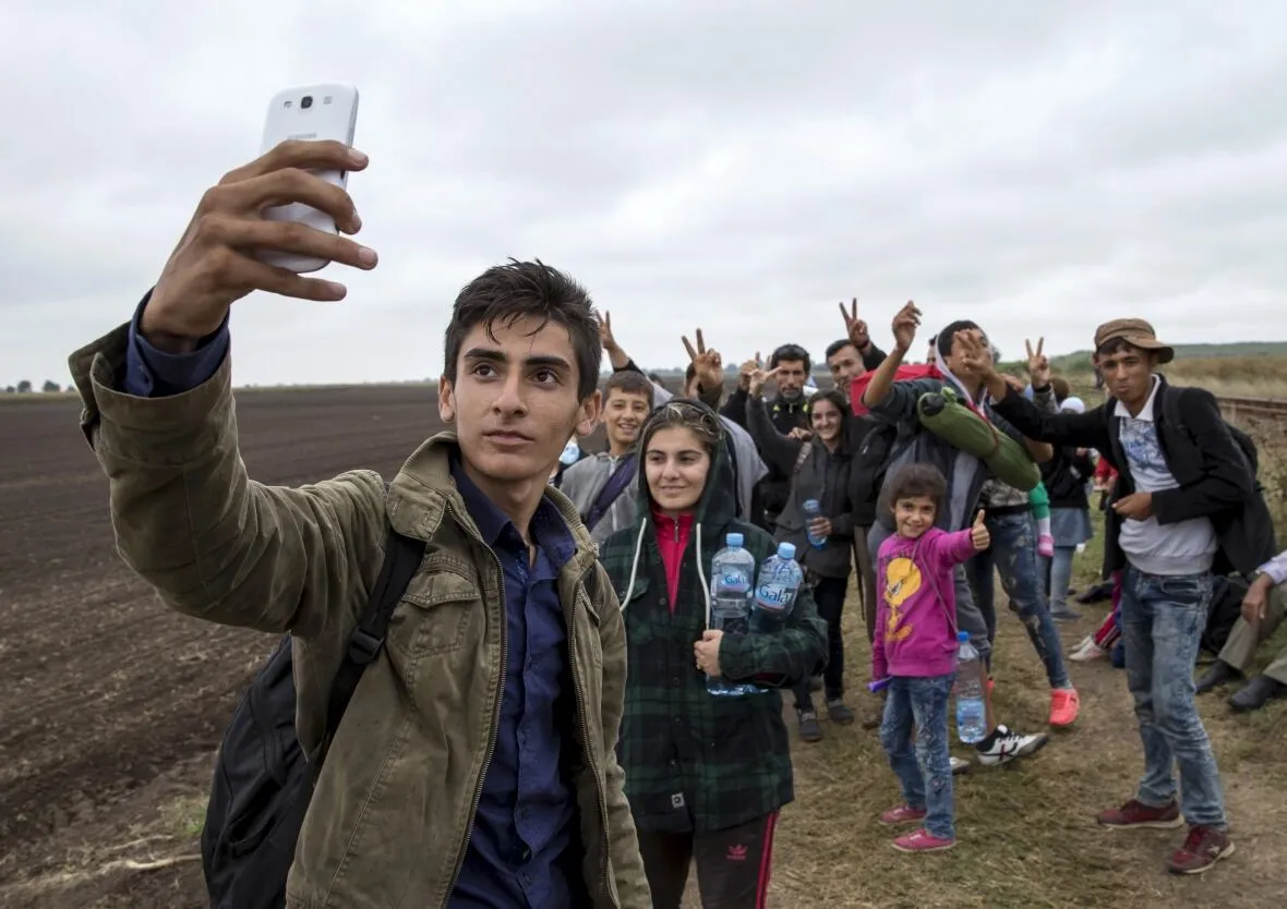 A young man takes a selfie with his phone on a railway track in open country, friends behind him raising peace signs.